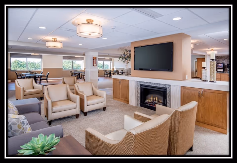 A spacious and well-lit common area in a senior living facility featuring a cozy seating arrangement with beige armchairs and a gray sofa around a fireplace with a mounted flat-screen TV above it. In the background, there are dining tables and chairs near large windows letting in natural light. The room has modern ceiling lights and neutral-toned walls and carpet.