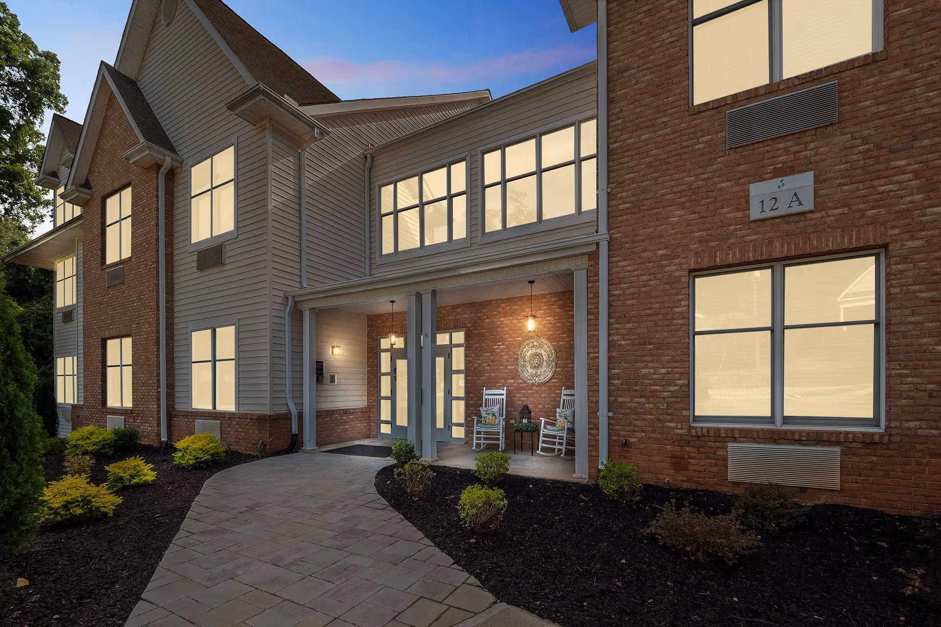 Exterior view of a two-story senior living facility building at dusk with a paved walkway leading to a covered entrance. The building features a combination of brick and siding, multiple windows, and a small porch area with two white rocking chairs and a decorative wall hanging. Landscaping includes small shrubs and mulch beds along the walkway.