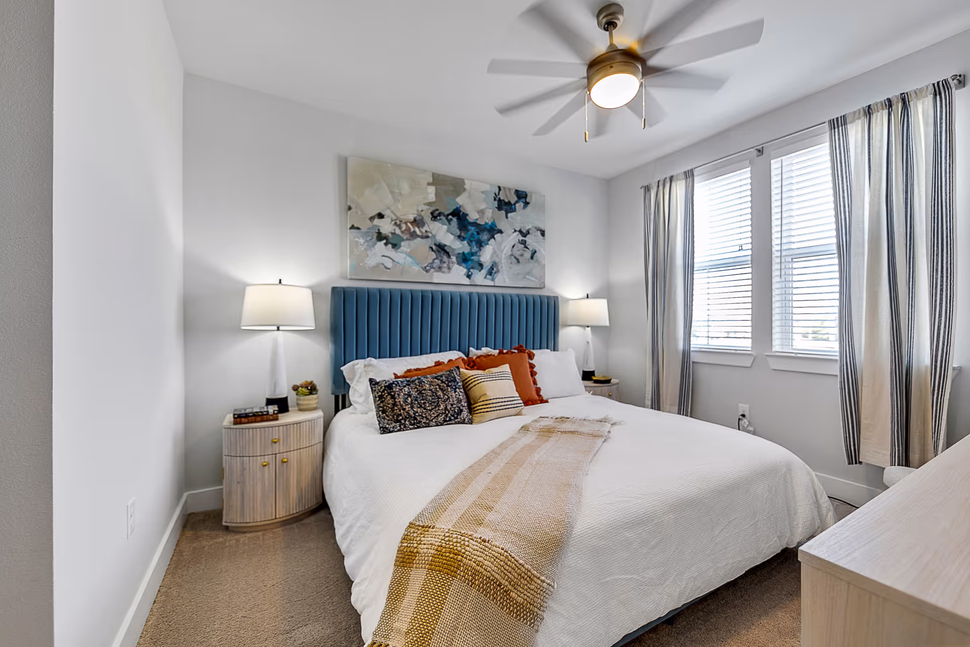 Well-lit bedroom with a bed featuring a blue upholstered headboard, bedside tables and lamps, a ceiling fan, and striped window curtains.