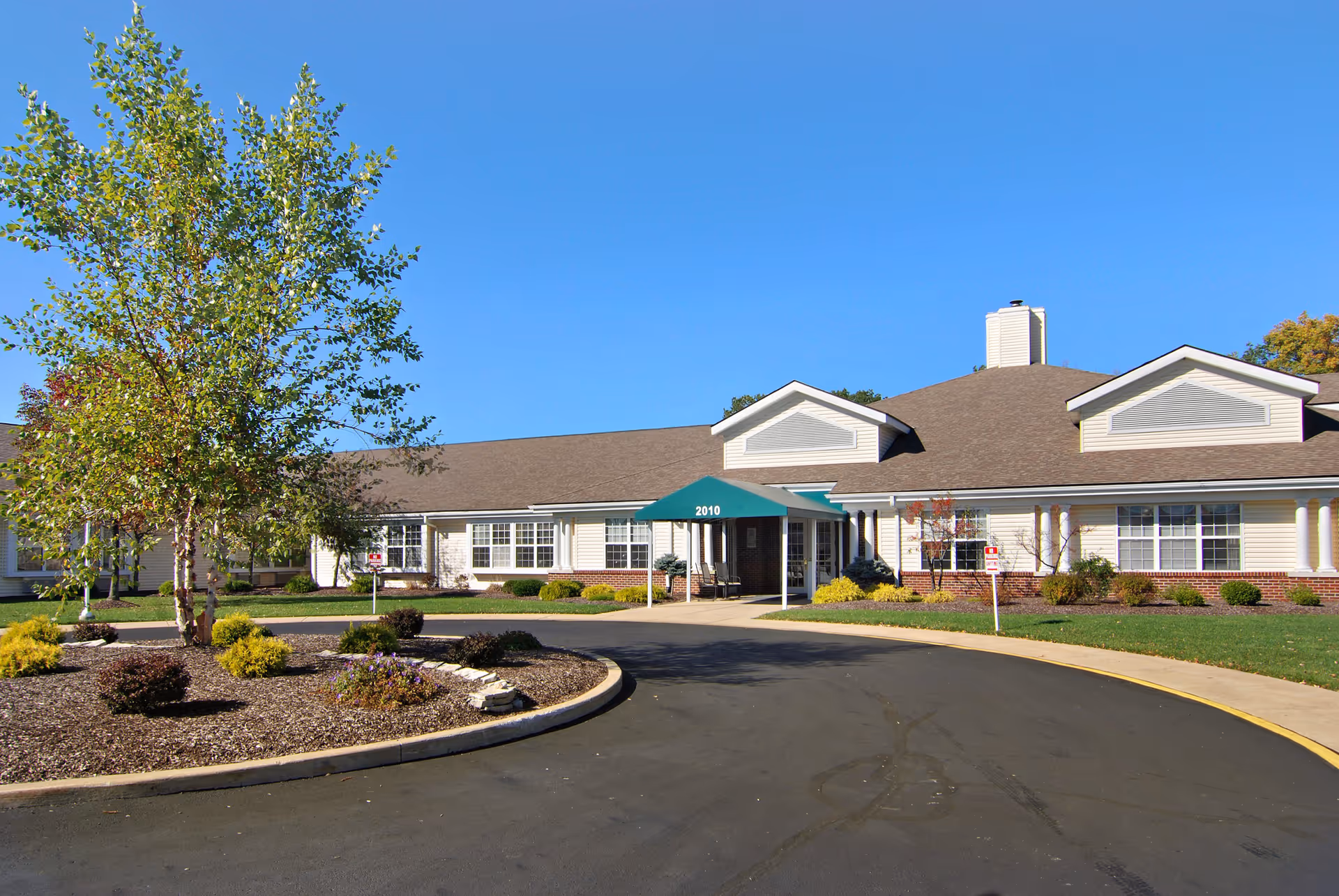 Front exterior view of Ontario Estates Senior Living facility with a curved driveway, landscaped garden with trees and shrubs, and a building entrance covered by a green awning displaying the number 2010.