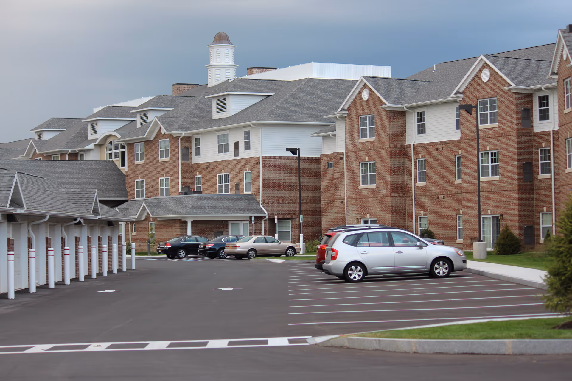 Exterior view of Towne Center Retirement Community showing a large brick building with multiple windows and a gray roof. There is a parking lot in the foreground with several parked cars and covered parking spaces on the left side. The sky is overcast.