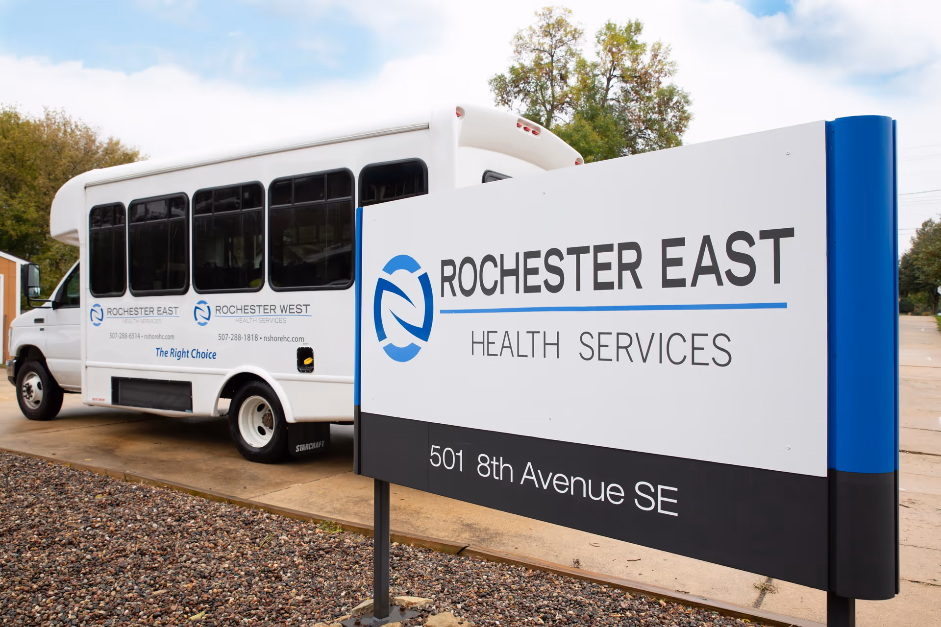 A white shuttle bus parked near a large sign for Rochester East Health Services located at 501 8th Avenue SE. The bus has logos and contact information for Rochester East and Rochester West Health Services. The area is outdoors with trees and a partly cloudy sky in the background.