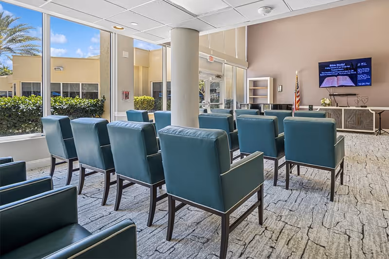 A bright room with large windows showing outdoor greenery, arranged with multiple teal upholstered chairs facing a wall-mounted TV screen displaying a Bible study announcement. The room has a carpeted floor, a column in the center, and an American flag near a cabinet and piano.