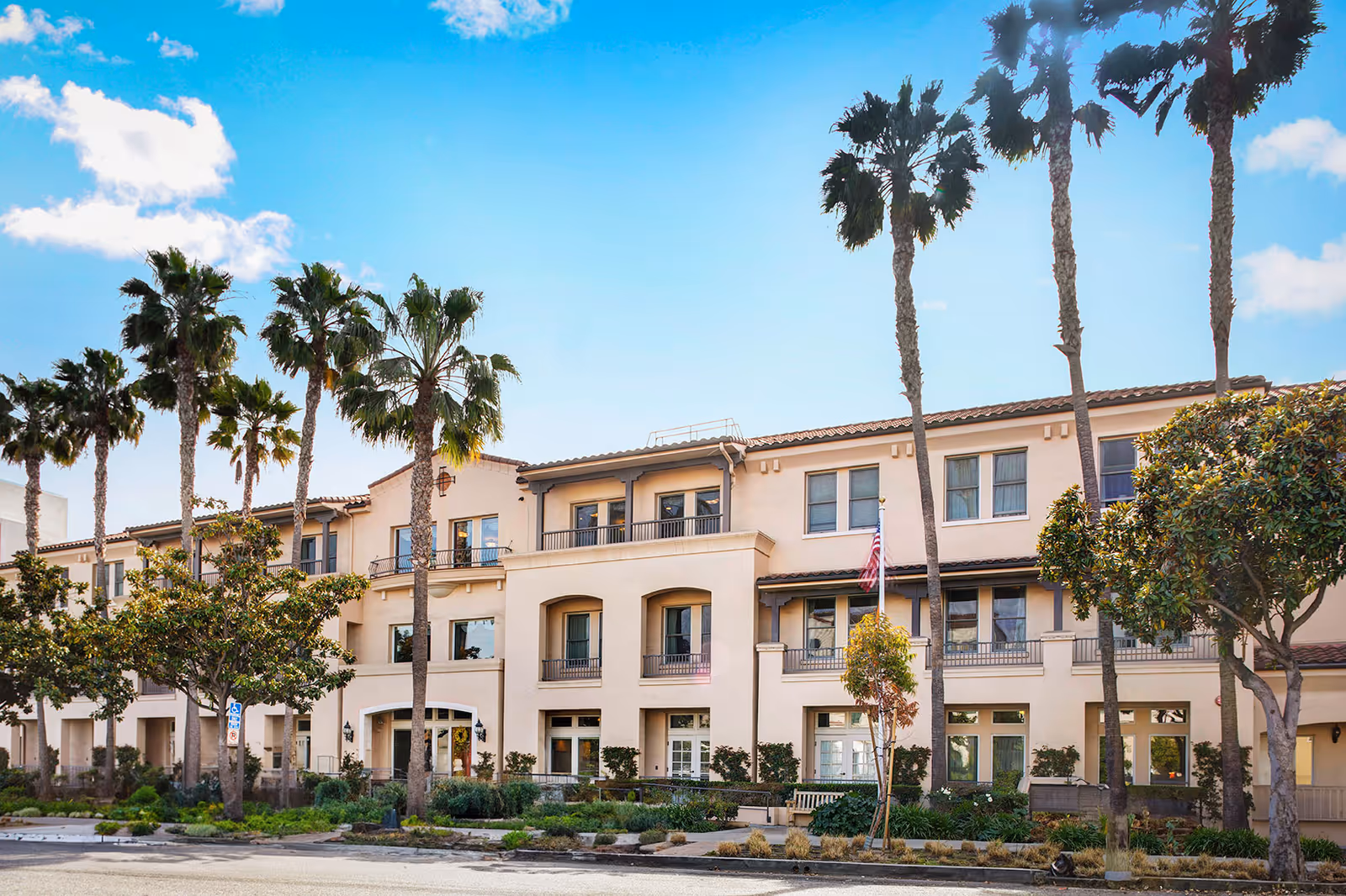 Three-story Mediterranean-style senior living building front with balconies, palm trees, and a clear blue sky.