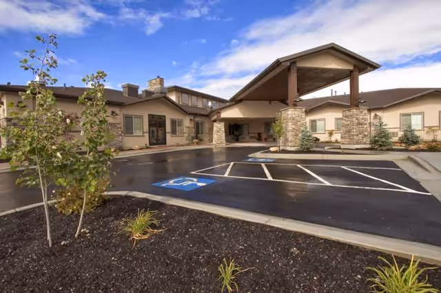 Exterior view of a single-story senior living facility building with a covered entrance, stone pillars, and a parking lot with a handicapped parking space. Small trees and plants are visible in landscaped areas near the building.