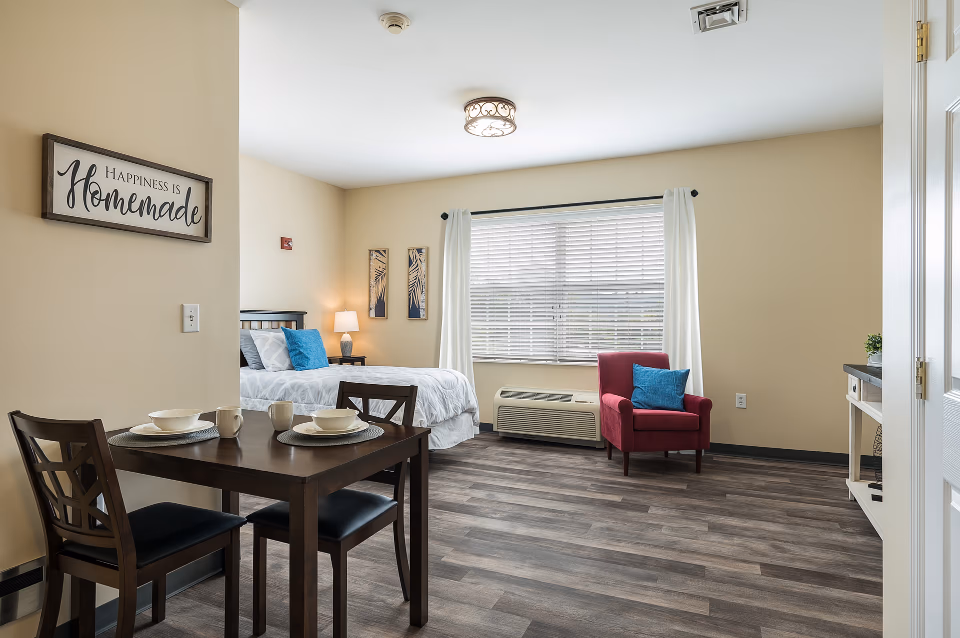 Bright studio apartment interior with a dining table in the foreground, a bed and bedside lamp, and a red armchair by a large window.