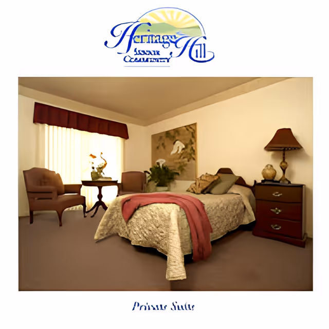 A cozy bedroom in a senior living facility with a bed covered in a beige quilt and a red throw blanket. There are two chairs and a small round table near a window with vertical blinds and a dark valance. A nightstand with a lamp and decorative items is beside the bed, and a framed artwork hangs on the wall.