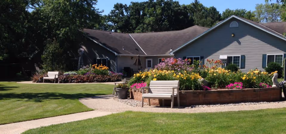 Front exterior of a single-story memory care building with colorful flower beds, benches, and a manicured lawn.