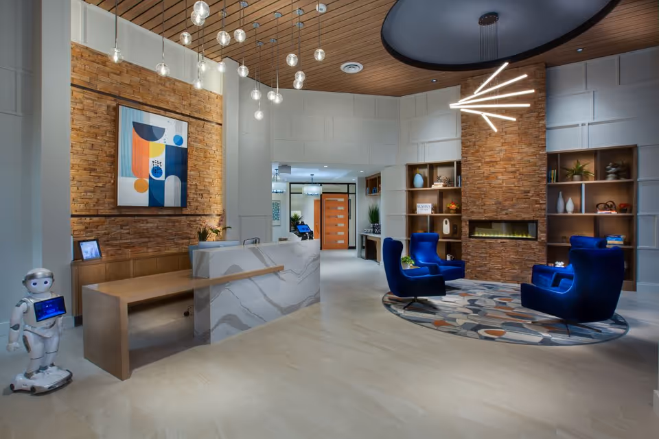Modern senior living facility lobby with a marble reception desk, wooden accent wall with abstract art, hanging pendant lights, a small humanoid robot on wheels, and a seating area featuring three blue armchairs arranged around a circular patterned rug in front of a stone fireplace with built-in shelves displaying decorative items.