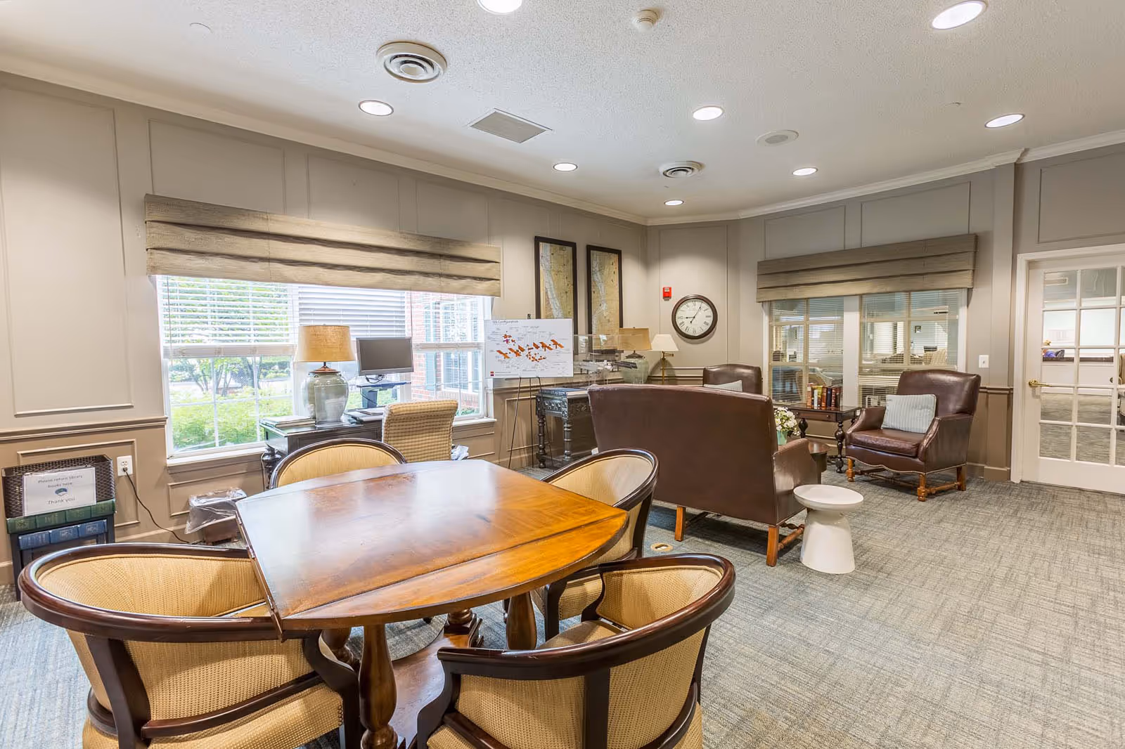 A bright senior living common room with a wooden table and chairs in the foreground and upholstered armchairs and a sofa near windows and a wall clock.