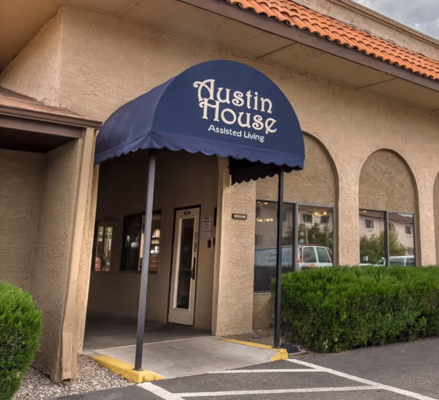 Entrance to Austin House Assisted Living with a blue awning over the doorway, beige stucco walls, arched windows, and green bushes along the building exterior.