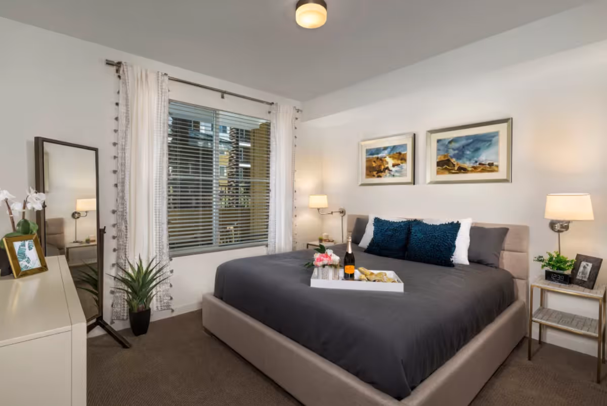 A well-styled bedroom with a large bed, bedside tables and lamps, a window with curtains, decorative artwork, and plants.