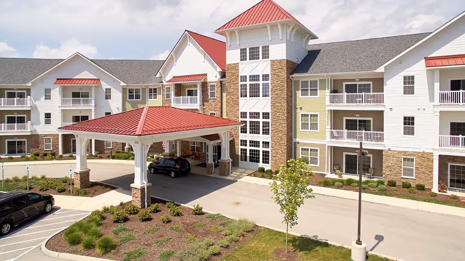 Exterior view of The Reserve at Hamilton Trace senior living facility showing a multi-story building with white and light green siding, stone accents, and red metal roofs. The entrance features a covered porte-cochère with cars parked nearby and landscaped greenery around the driveway.