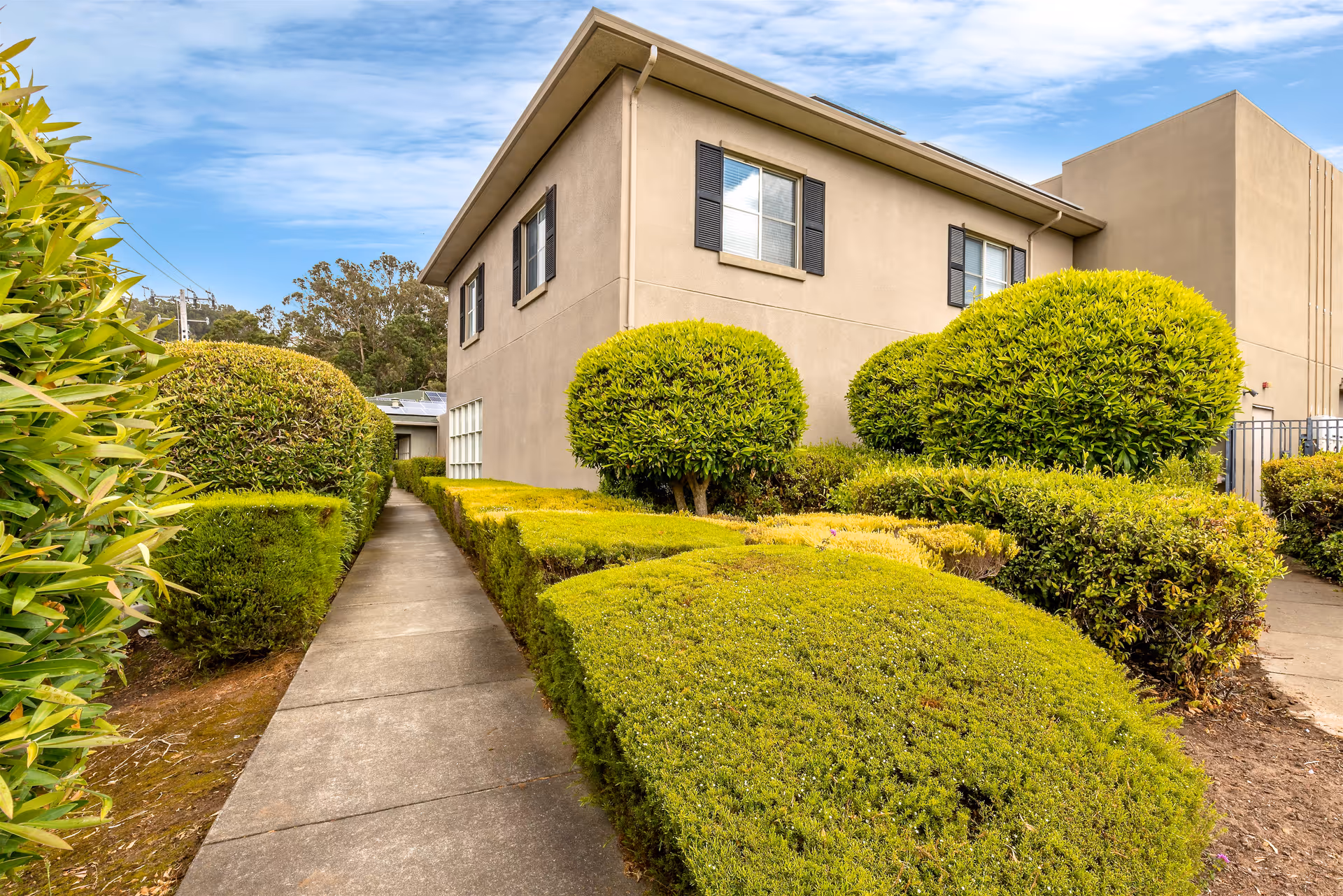 Side view of a beige two-story building with black window shutters, surrounded by neatly trimmed green bushes and a concrete walkway leading alongside the building under a partly cloudy blue sky.