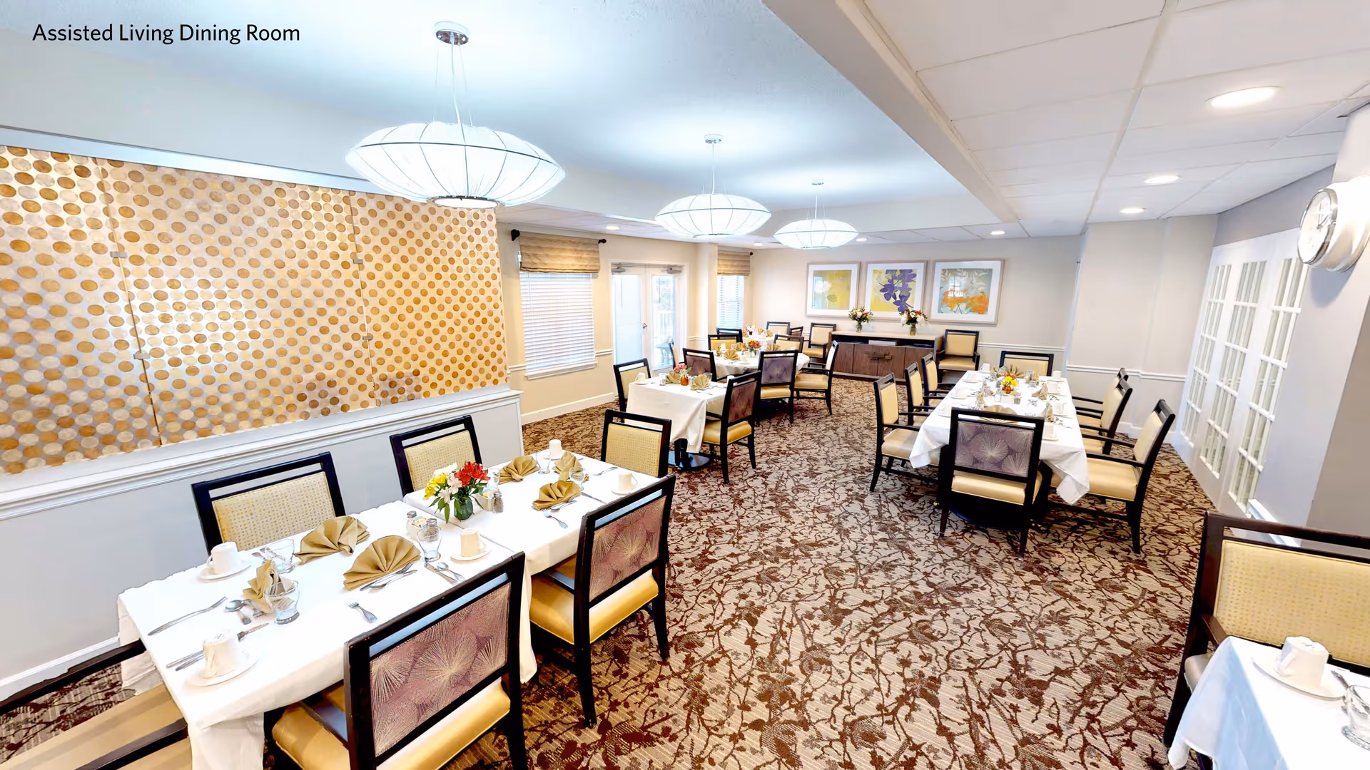 A bright assisted living dining room with multiple tables covered in white tablecloths, each set with beige napkins, cups, glasses, and silverware. The room features patterned carpet, modern hanging light fixtures, framed floral artwork on the walls, and a decorative wall panel with a circular pattern. There are windows with blinds and a door leading outside.