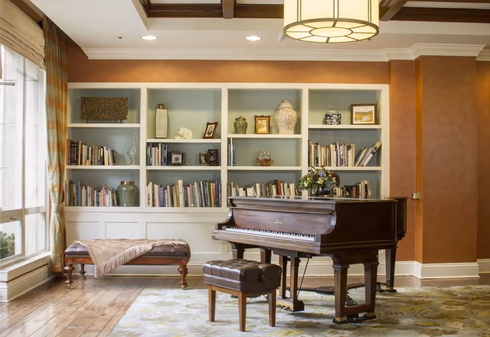 A cozy living room area featuring a grand piano with a matching piano bench, a cushioned bench with a throw blanket, and built-in white bookshelves filled with books and decorative items. The room has warm brown walls, wooden flooring, a large window with curtains, and a hanging ceiling light fixture.
