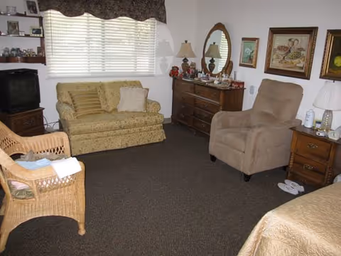 Senior living bedroom with a loveseat, recliner, wicker chair, dresser with mirror, TV and a partially visible bed.
