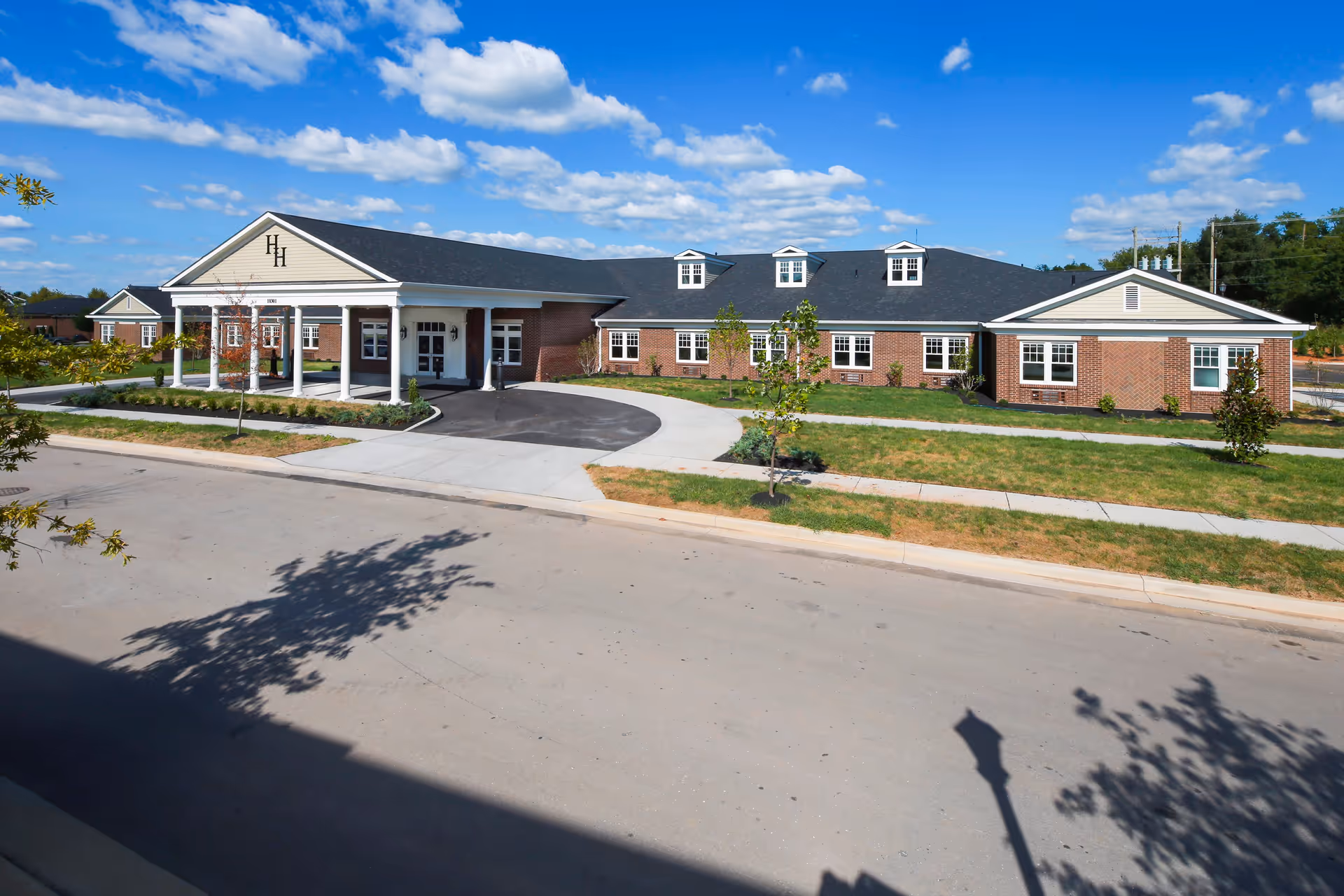Exterior view of a single-story brick building with a covered entrance supported by white columns, a curved driveway, and a well-maintained lawn under a blue sky with scattered clouds. The building has a dark shingled roof with three dormer windows and the letters 'HH' displayed on the front gable.