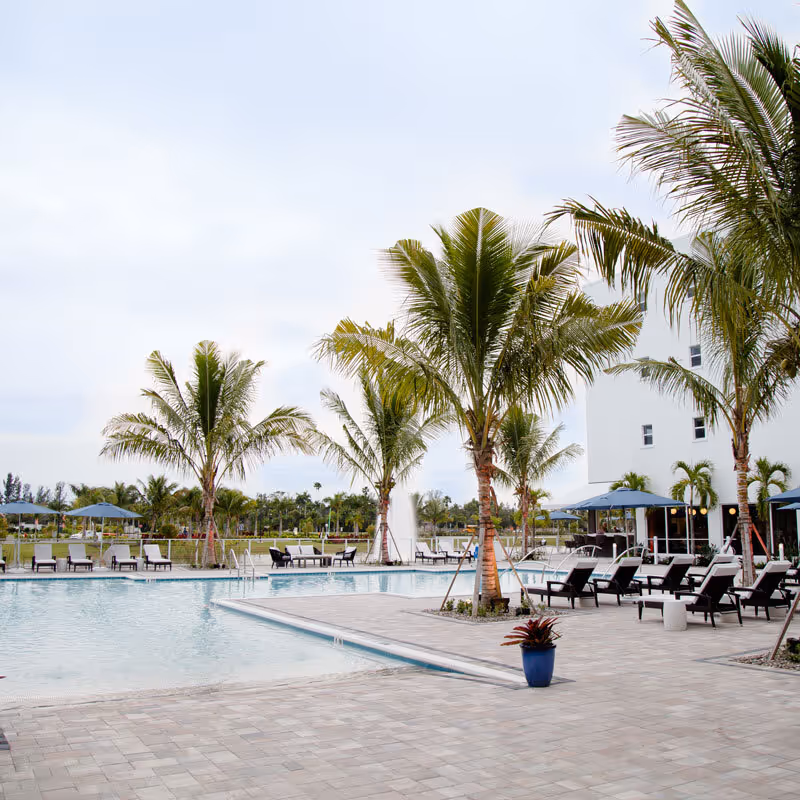 Outdoor swimming pool area with lounge chairs and umbrellas surrounded by palm trees at a senior living facility. A white multi-story building is visible in the background along with a water fountain and green landscaping.