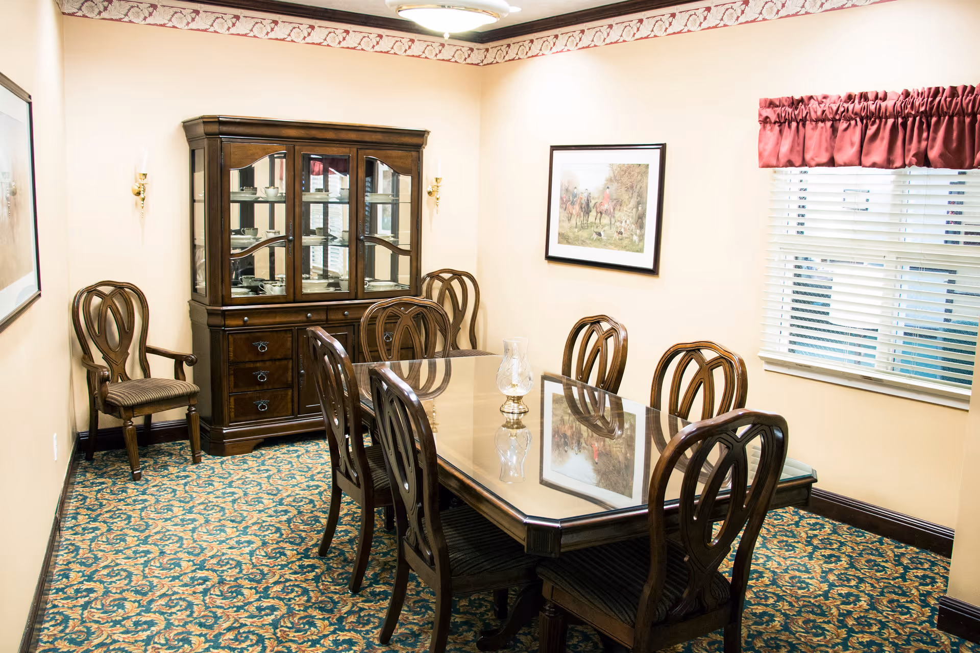 A traditional dining room featuring a glass-topped wooden table surrounded by ornate wooden chairs, a china cabinet, framed art, and a window with blinds.