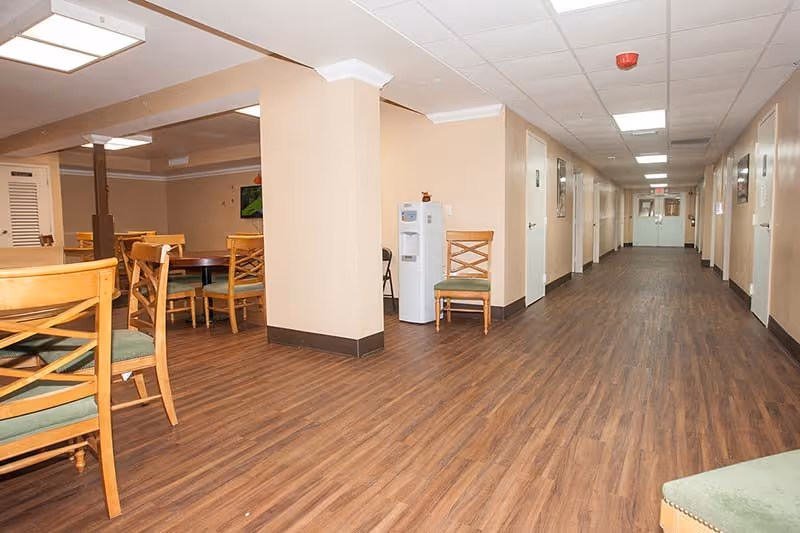 Interior view of a senior living facility hallway with wood flooring and beige walls. On the left side, there is a dining area with wooden chairs and tables. A water cooler and a single chair are positioned near a column. The hallway extends to double doors at the far end, with several closed doors along the right side.