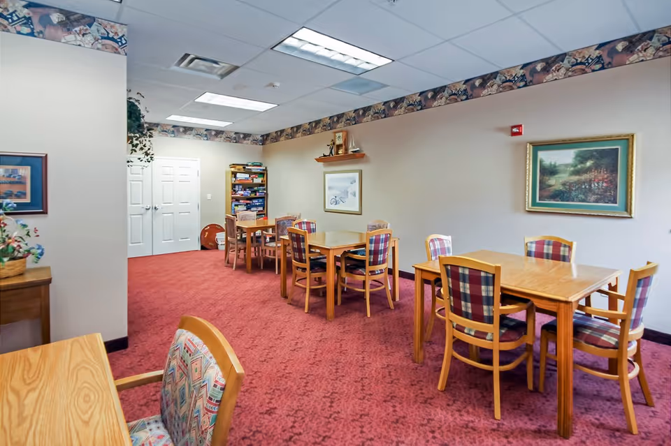 A common area with multiple wooden tables and chairs with plaid upholstery on a red carpeted floor. The walls are beige with framed artwork and a decorative border near the ceiling. There is a bookshelf filled with board games and other items in the background, and a small table with a plant on the left side.
