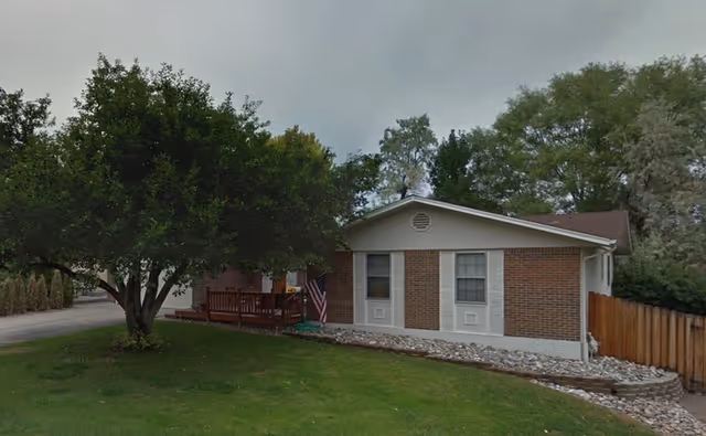 Exterior view of a single-story brick building with white trim, a small wooden porch with an American flag, surrounded by green grass and trees under a cloudy sky.
