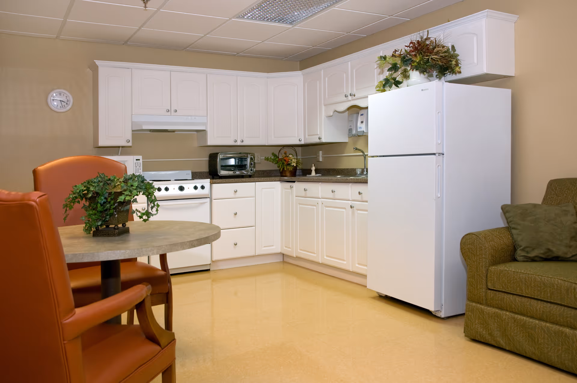 A compact kitchenette and seating area with white cabinets, a stove and refrigerator, a round table with chairs, and a sofa.