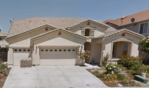 Front exterior view of a beige stucco house with a tiled roof, three garage doors, a driveway, and a small garden with flowers and shrubs.