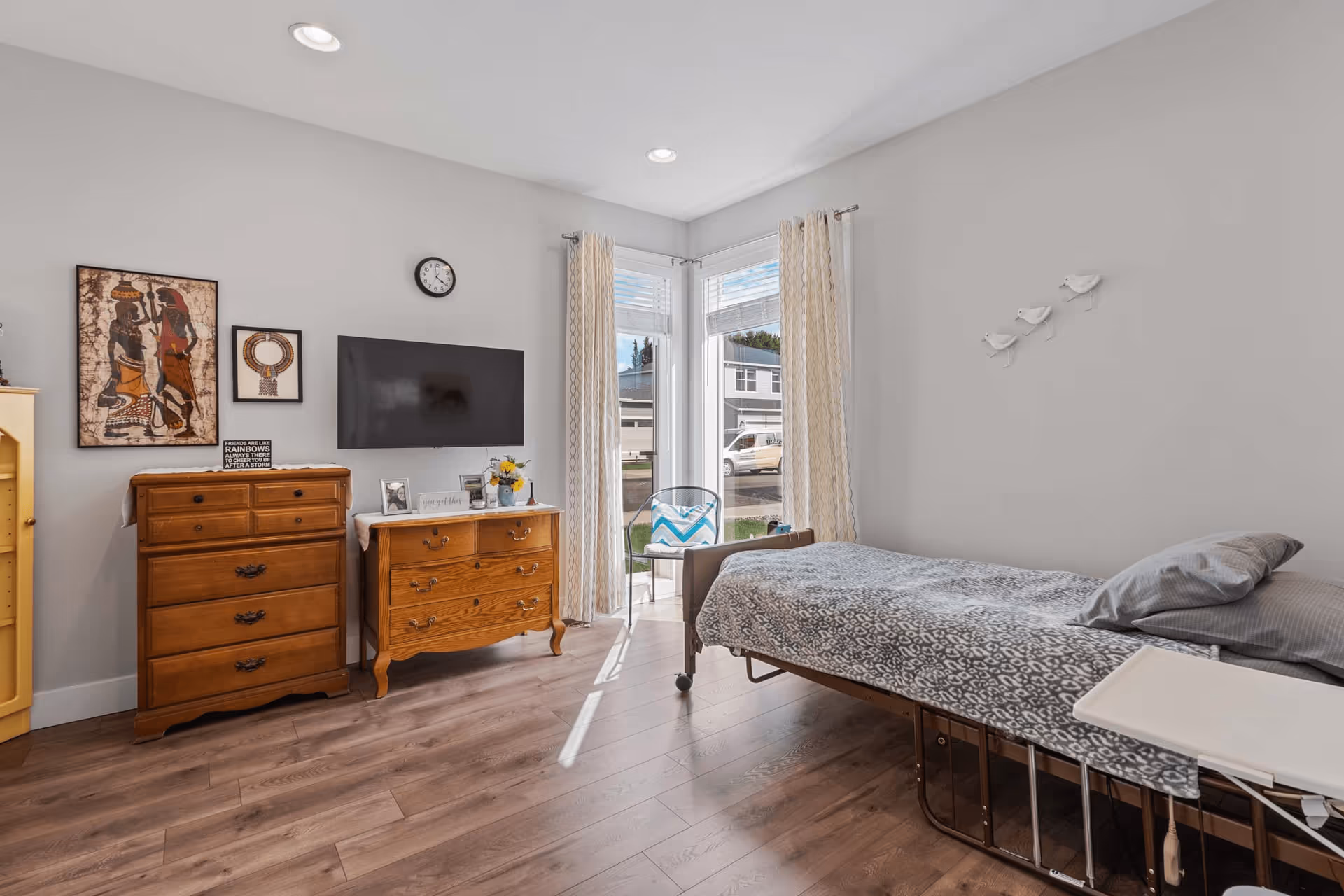 A bright and clean bedroom in a senior living facility featuring a single bed with gray patterned bedding and pillows, a small white overbed table, two wooden dressers with framed photos and decorative items, a wall-mounted flat screen TV, a clock, and two large windows with sheer curtains letting in natural light. The room has light gray walls, wood flooring, and some wall art including three small white bird decorations.