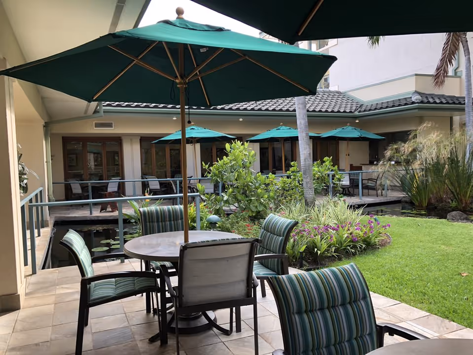 Outdoor patio area at Kāhala Nui with round tables and striped cushioned chairs under green umbrellas. The patio overlooks a small garden with green grass, plants, and flowers, and is adjacent to a building with large windows and a tiled roof.