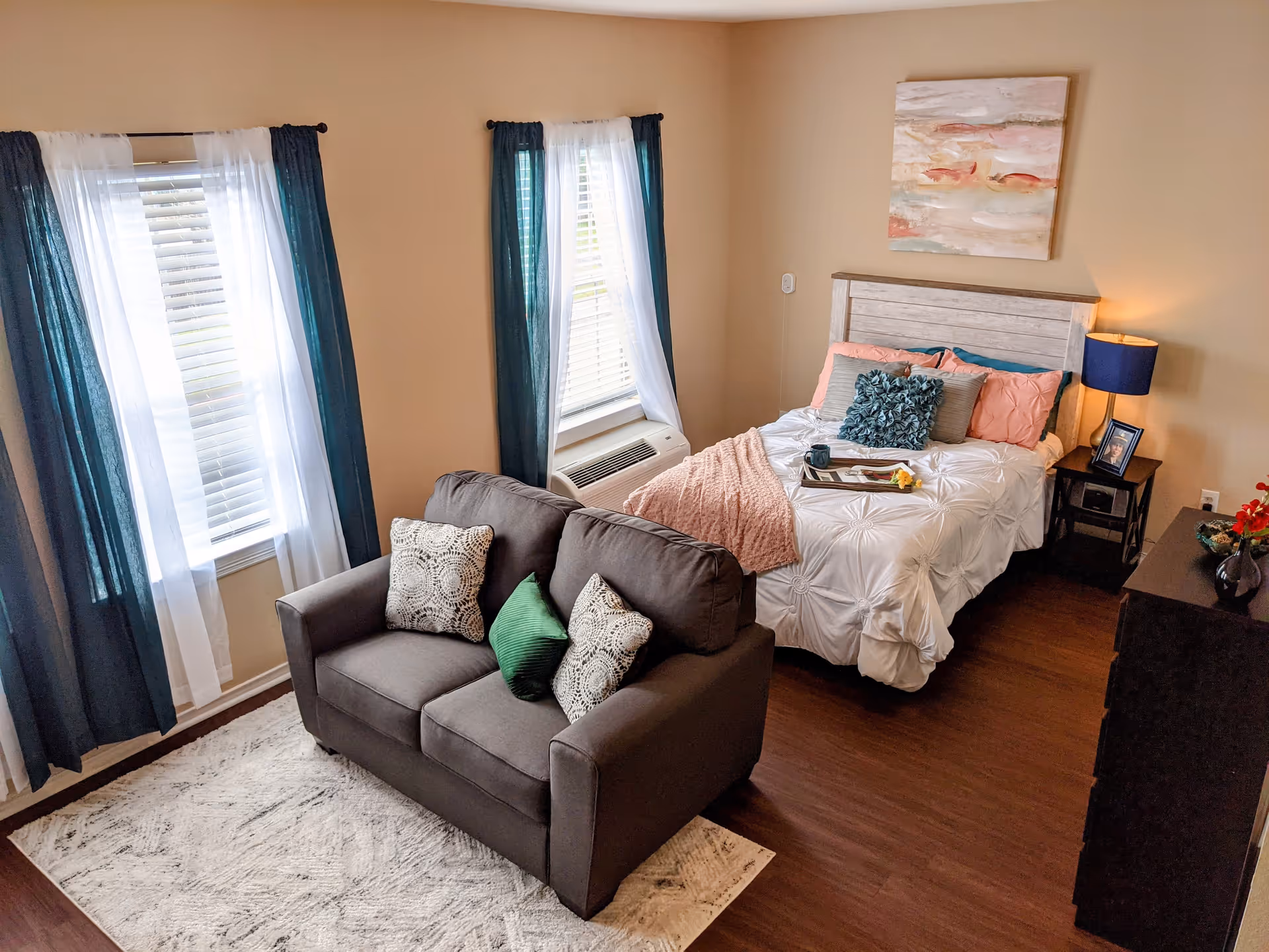 A cozy bedroom with a bed dressed in white bedding and multiple pillows, a small gray loveseat with decorative pillows, two windows with white and dark teal curtains, a nightstand with a blue lamp and a framed photo, a dresser with a vase of flowers, and a piece of abstract wall art above the bed.