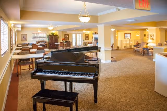 Interior view of a spacious common area in a senior living facility featuring a black grand piano with a matching bench in the foreground. The room has beige walls, carpeted floors, multiple tables and chairs, and warm lighting from ceiling fixtures. There are windows with blinds on the left side and various pieces of furniture and decor in the background.