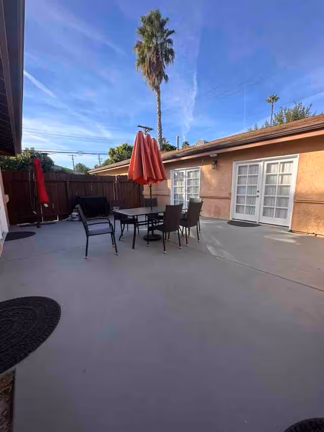 Outdoor patio area with a table and four chairs, two red umbrellas, a barbecue grill, and a tall palm tree in the background under a clear blue sky. The patio is surrounded by a wooden fence and adjacent to a building with beige walls and white French doors.