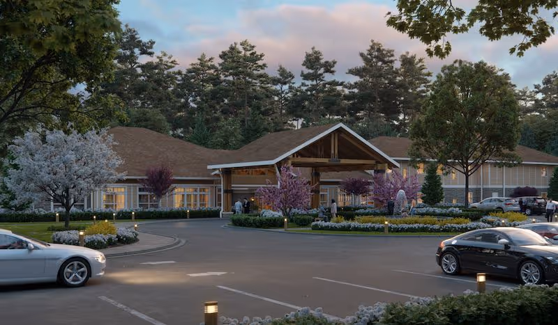Exterior view of Prema at Ashton Hills senior living facility with a circular driveway, landscaped garden with trees and flowering plants, several parked cars, and people near the entrance under a wooden canopy roof during early evening.