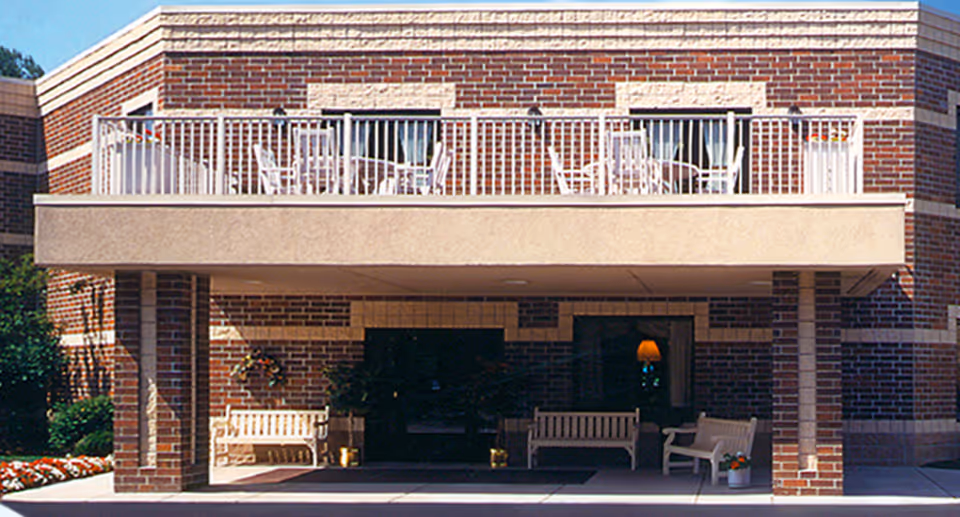 Front exterior of a brick assisted living building with a covered entrance, white benches, and an upper balcony with white railings and chairs.