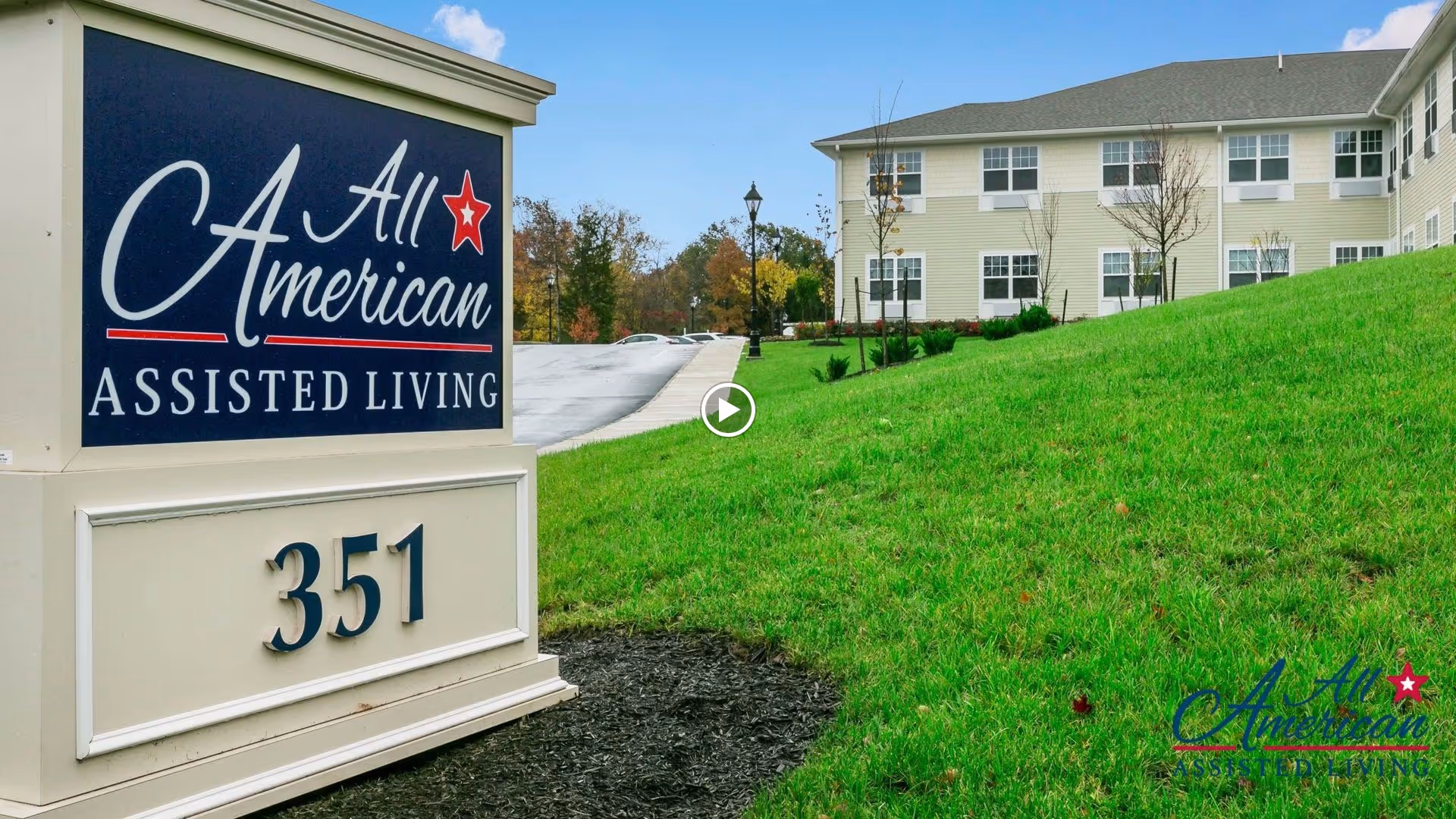 Entrance sign reading All American Assisted Living and 351 beside a grassy lawn with the facility building in the background.