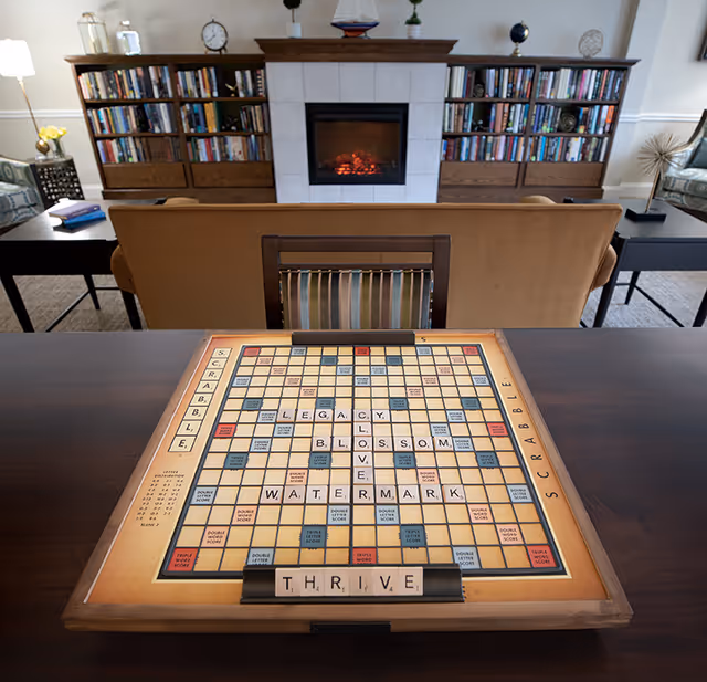 A cozy living room with a Scrabble board on a wooden table in the foreground. The Scrabble board has words like LEGACY, BLOSSOM, LOVE, WATERMARK, and THRIVE spelled out. Behind the table is a brown sofa facing a fireplace with a glowing fire. On either side of the fireplace are wooden bookshelves filled with books and decorative items. The room is warmly lit and has a comfortable, inviting atmosphere.