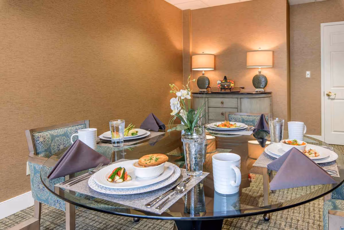 A dining room with a round glass table set for four people. Each place setting includes a plate with food, a folded dark napkin, a glass of water, and a white mug. The chairs have patterned upholstery. In the background, there is a sideboard with two lamps and decorative items, and a closed white door on the right.