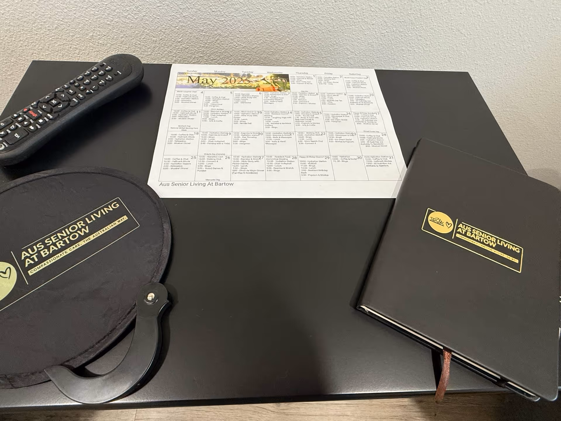 A black table with a remote control, a folded black fan with the logo and text 'AUS SENIOR LIVING AT BARTOW Compassionate Care. The Australian Way.', a black notebook with the same logo and text, and a printed calendar for May 2025 from Aus Senior Living At Bartow.