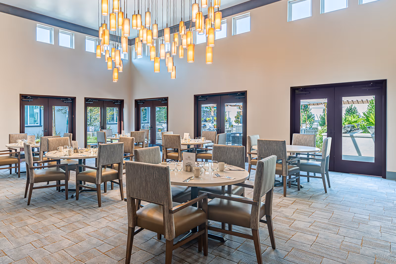 A bright dining room with multiple round tables and cushioned chairs arranged neatly. The room features large windows and glass doors letting in natural light, with a modern chandelier hanging from the high ceiling. The tables are set with cups, glasses, and silverware, ready for a meal.