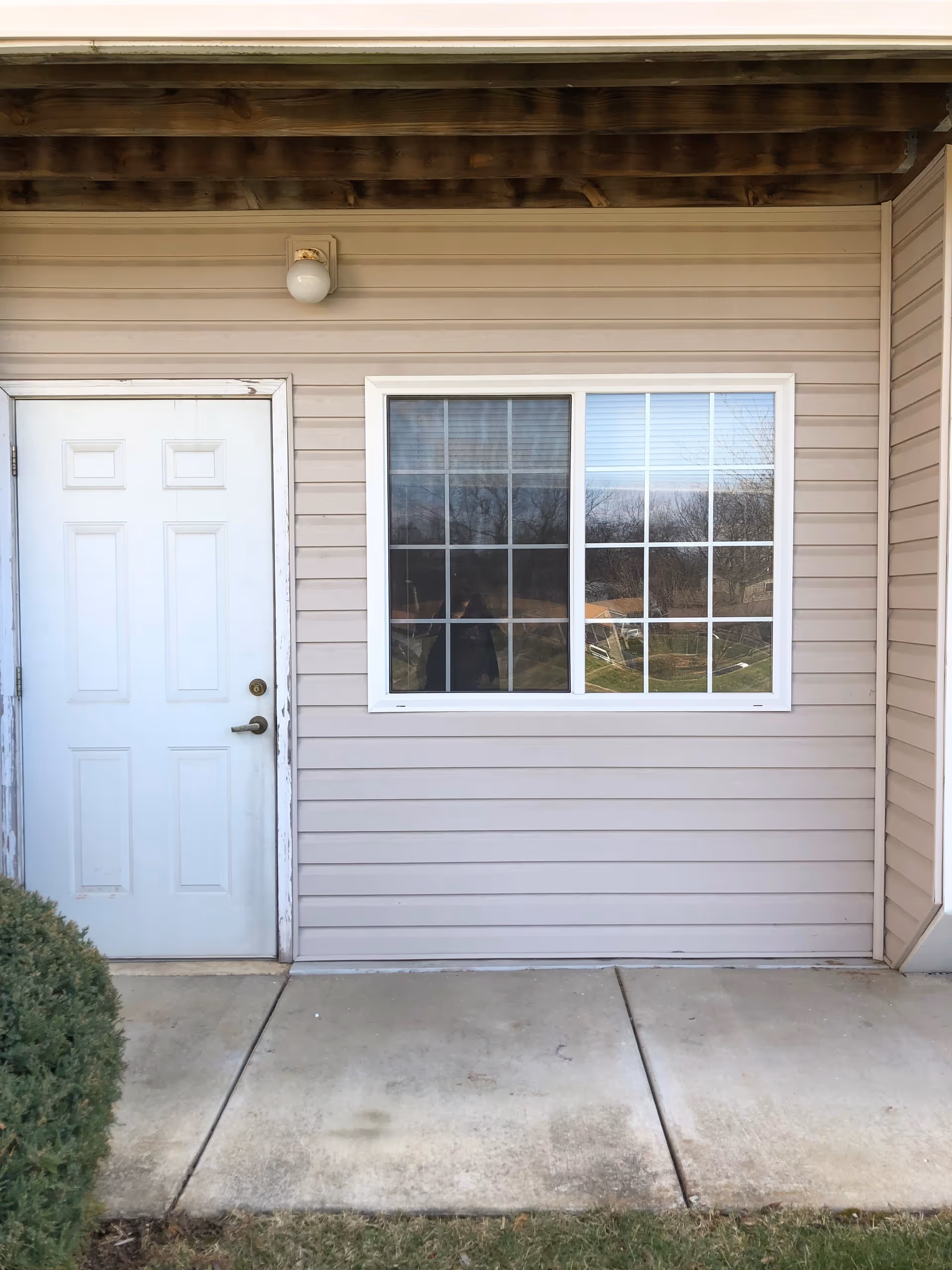 Exterior view of a beige building wall with a white door on the left and a window with white grid panes on the right. There is a small bush in the bottom left corner and a concrete walkway in front of the building. A light fixture is mounted above the door.