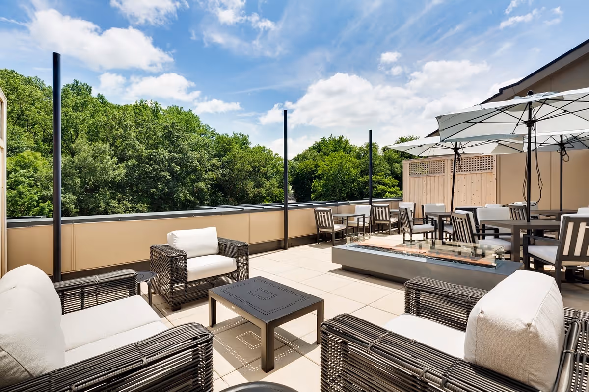 Outdoor patio area with cushioned wicker chairs, tables, large umbrellas, and a modern fire pit, surrounded by a railing and overlooking green trees under a partly cloudy blue sky.