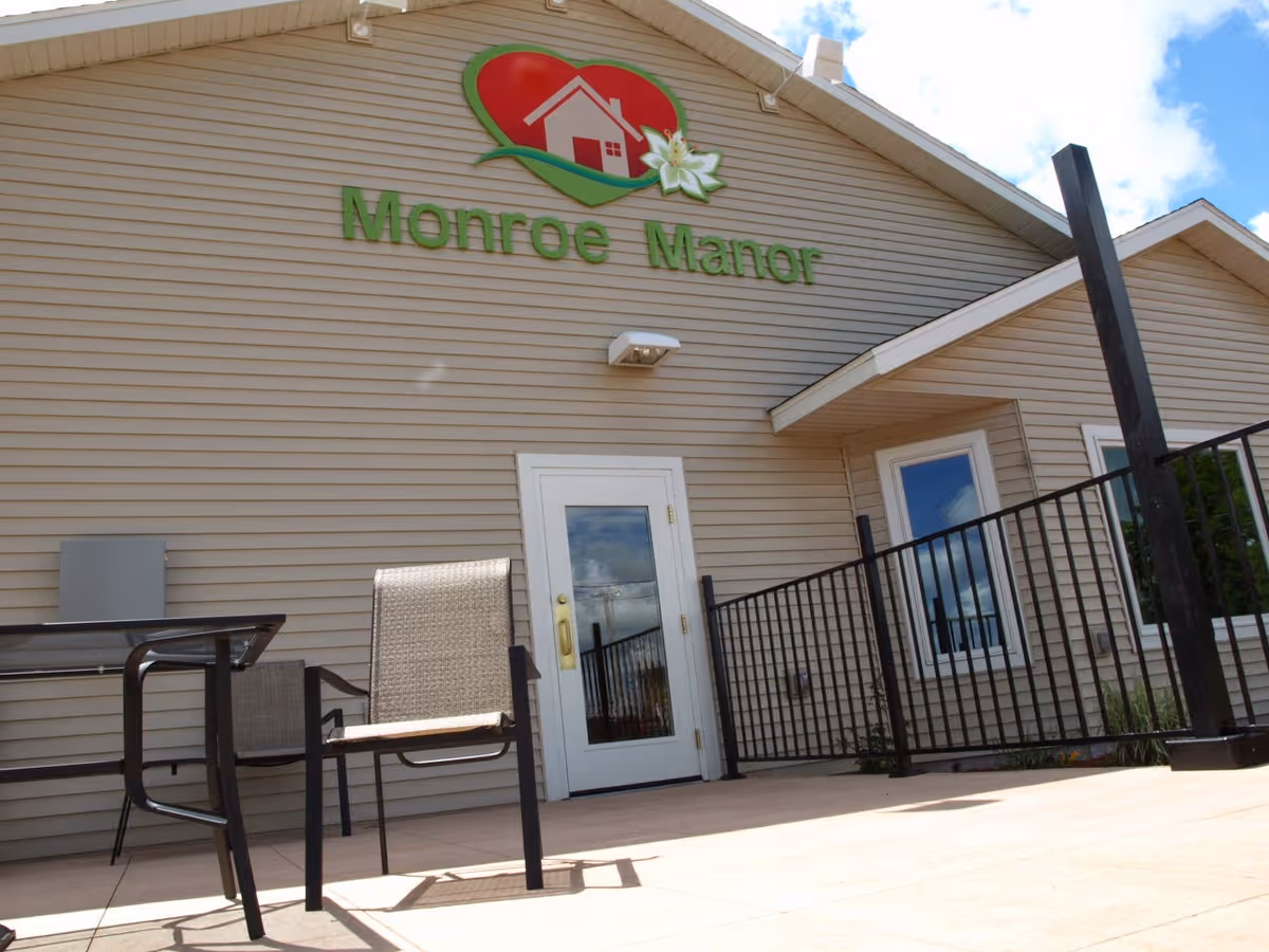Front entrance of Monroe Manor showing the building's sign above a glass door, a small patio with chairs, and a railing.