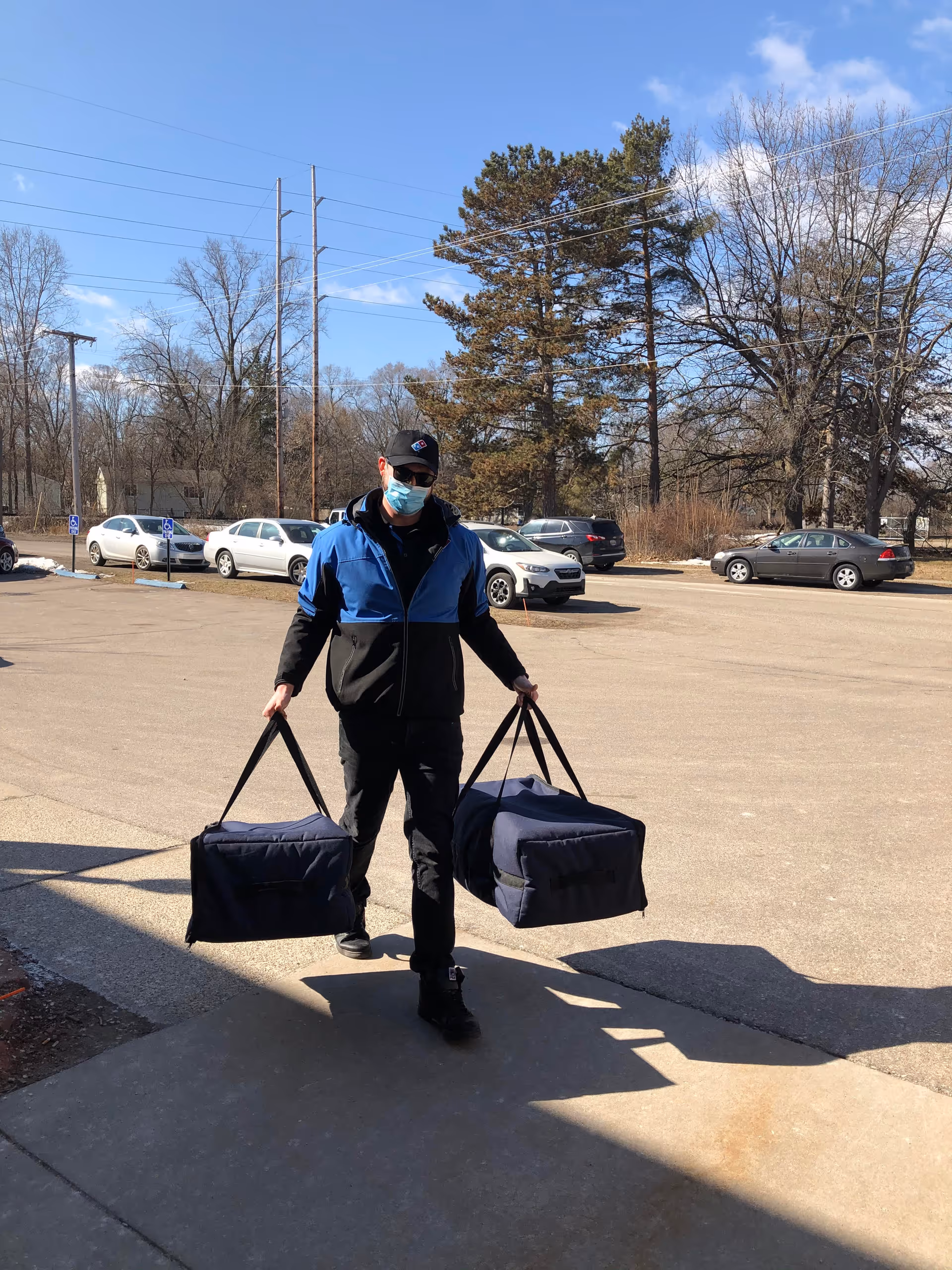 A man wearing a blue and black jacket, black pants, a black cap, sunglasses, and a face mask is walking outside on a sunny day carrying two large insulated delivery bags, one in each hand. There are several parked cars and leafless trees in the background under a clear blue sky.