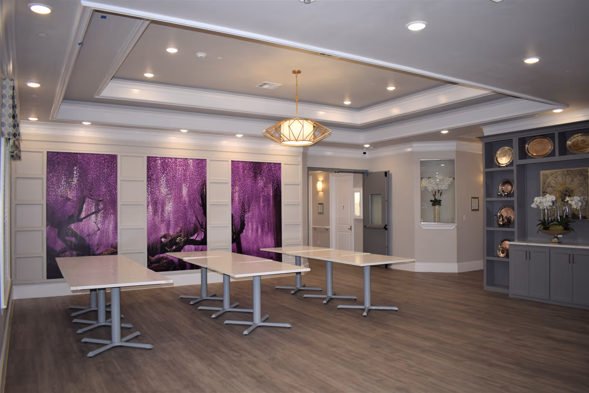 A spacious, well-lit interior room with three rectangular tables arranged in a U-shape. The room features a tray ceiling with recessed lighting and a modern pendant light fixture. The back wall has three large panels with purple floral artwork. To the right, there is a built-in gray cabinet with decorative plates and white orchids. The floor is wood-style, and there is a door and a small windowed alcove on the far wall.