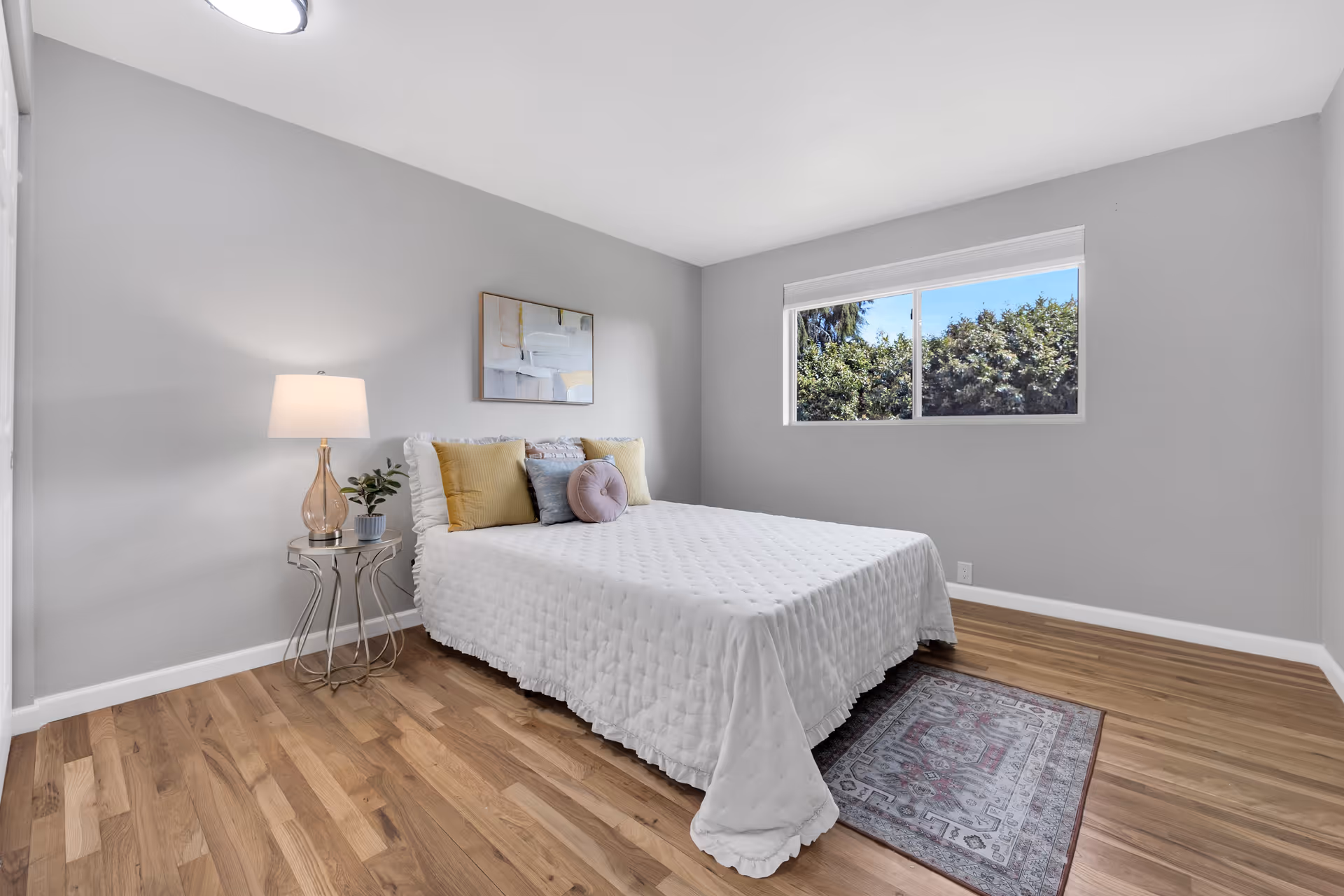 A bedroom with a neatly made bed covered in a white quilt and decorated with several pillows in yellow, gray, and lavender. There is a small side table with a lamp and a plant next to the bed. A window on the right wall shows green trees outside. The room has light gray walls, wooden flooring, and a small rug partially under the bed.