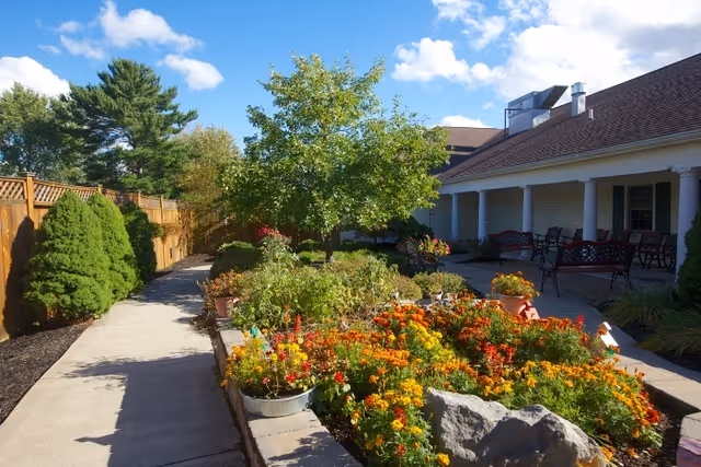 Sunny courtyard garden with colorful flower beds, a tree, a paved walkway and benches along a covered porch.