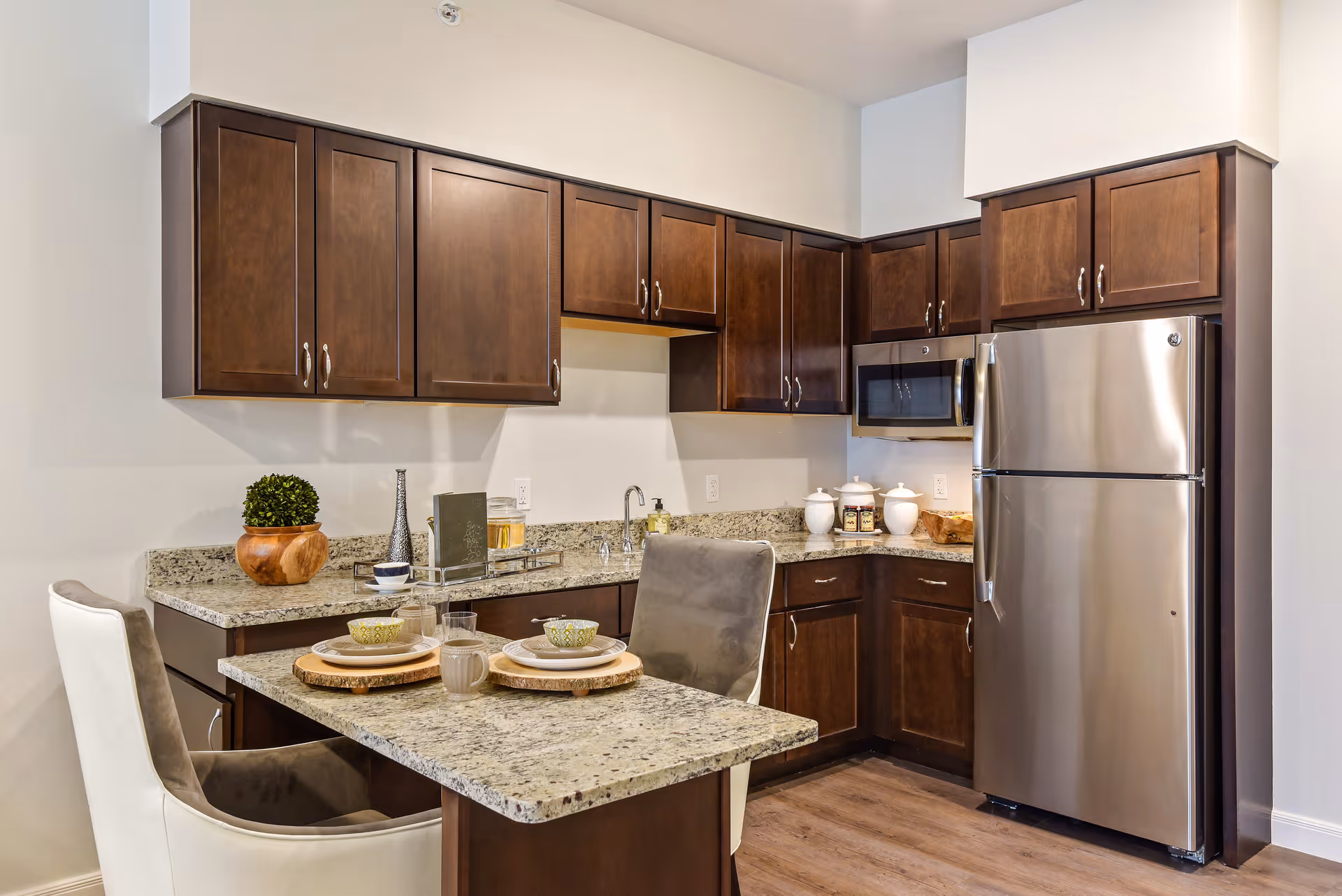Modern kitchen with dark wood cabinets, granite countertops, stainless steel refrigerator and microwave, and a small dining area with two chairs and place settings.