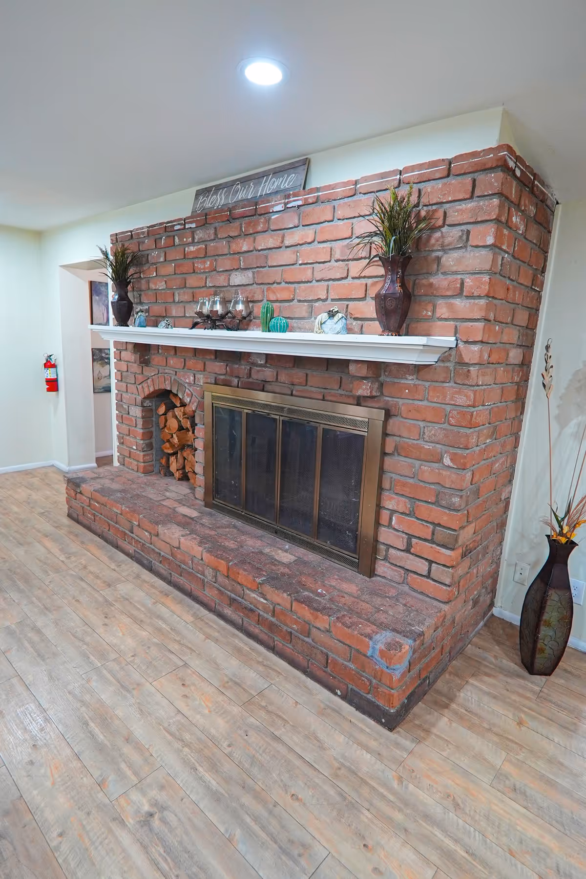 A cozy brick fireplace with a brass screen in a room with light wood flooring. The fireplace mantel is decorated with two vases containing plants, candles, and small decorative items. A sign above the mantel reads 'Bless Our Home'. There is a fire extinguisher mounted on the wall in the background.